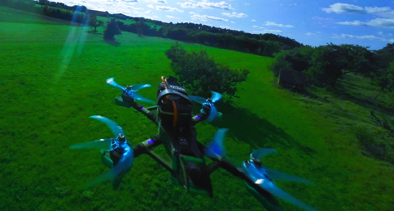 Das Bild zeigt einen Quadcopter mit blauen propellern und blauen LEDs an den Armen beim Flug über einer grünen Obstbaumwiese. der Himmel ist aufgelockert und die Sonne scheint.