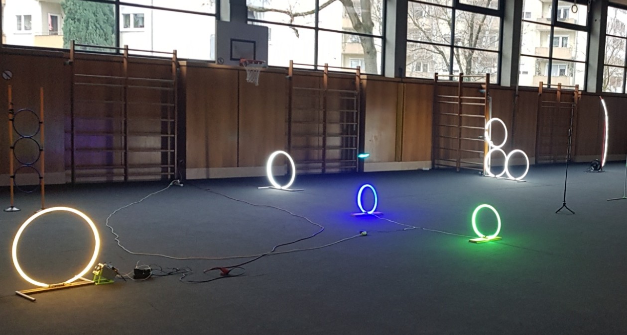 Das Bild zeigt einen Parcours mit runden LED-Gates in einer Sporthalle mit dunklem Boden und großen Fenstern. Es befinden sich Kletterwände an der Seite.